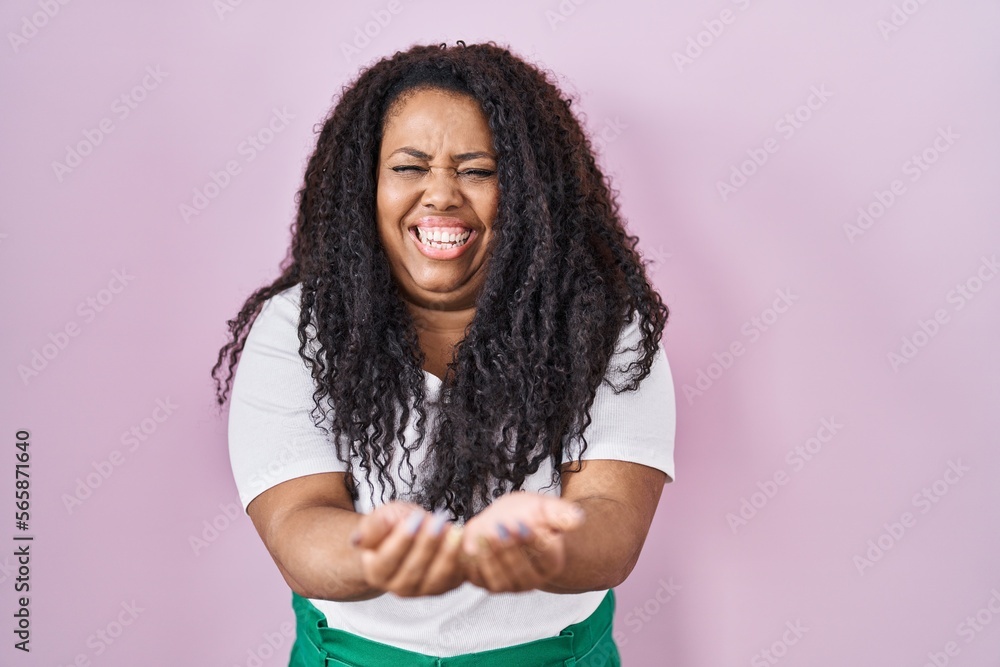 Plus size hispanic woman standing over pink background smiling with hands palms together receiving or giving gesture. hold and protection Plus size hispanic woman standing over pink background smiling with hands palms together receiving or giving gesture. hold and protection
