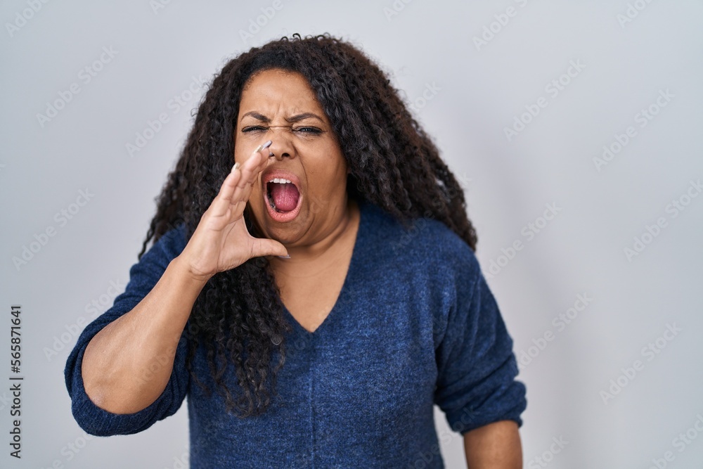 Plus size hispanic woman standing over white background shouting and screaming loud to side with hand on mouth. communication concept. Plus size hispanic woman standing over white background shouting and screaming loud to side with hand on mouth. communication concept.