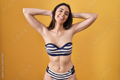 Young brunette woman wearing bikini over yellow background relaxing and stretching, arms and hands behind head and neck smiling happy