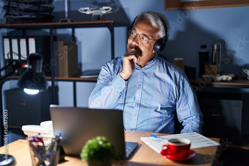Hispanic senior man wearing call center agent headset at night with hand on chin thinking about question, pensive expression. smiling with thoughtful face. doubt concept.