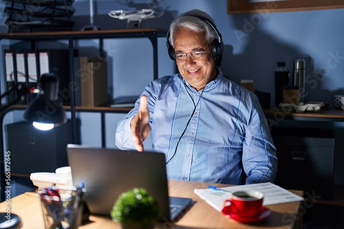 Hispanic senior man wearing call center agent headset at night smiling friendly offering handshake as greeting and welcoming. successful business.