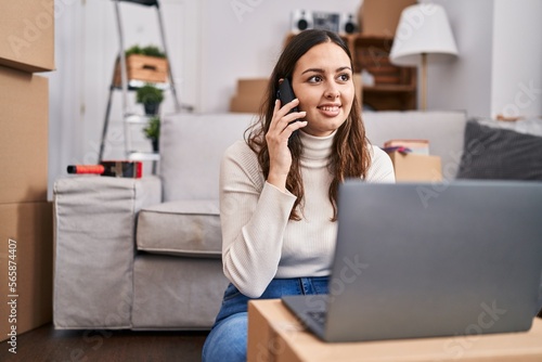 Young beautiful hispanic woman using laptop talking on smartphone at new home