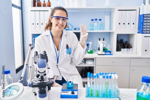 Young hispanic woman working at scientist laboratory showing and pointing up with fingers number three while smiling confident and happy.