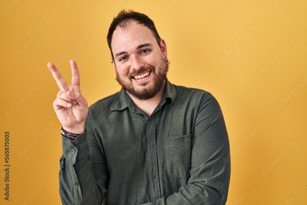 Plus size hispanic man with beard standing over yellow background smiling with happy face winking at the camera doing victory sign with fingers. number two. Plus size hispanic man with beard standing over yellow background smiling with happy face winking at the camera doing victory sign with fingers. number two.