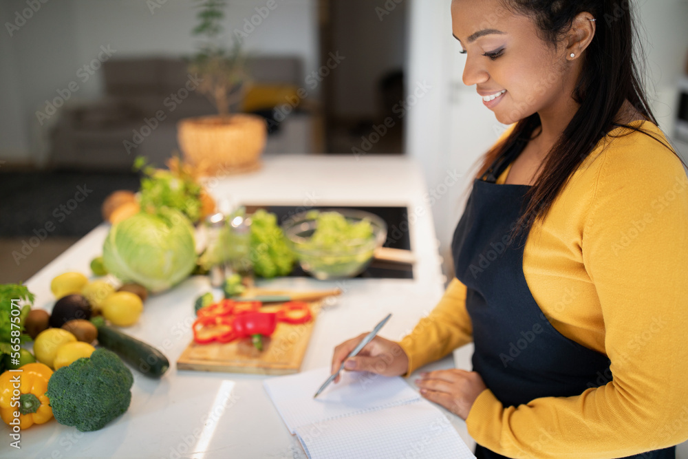 Glad millennial african american lady in apron make notes in notebook, read new recipe at book