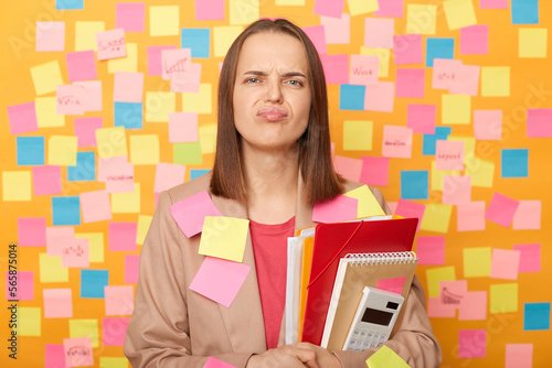 Image of sad upset woman wearing beige jacket posing over sticky notes to write reminder on yellow wall, holding paper documents, expressing sorrow and sadness, having problems with business.