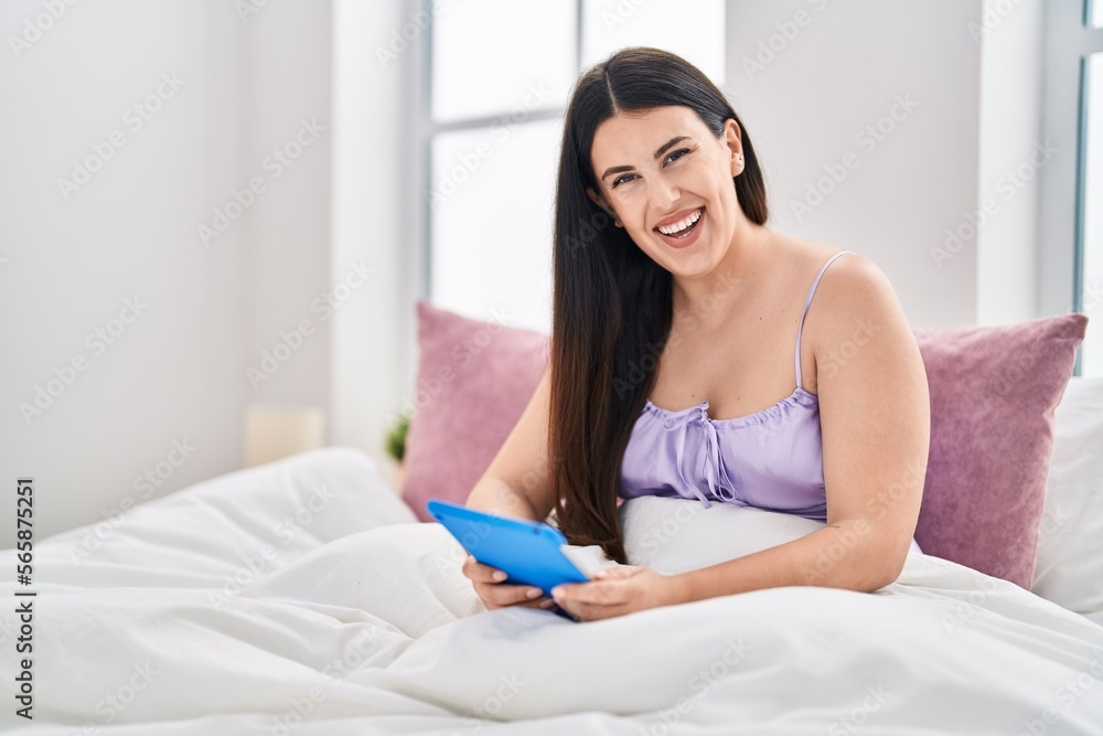 Young hispanic woman using touchpad sitting on bed at bedroom