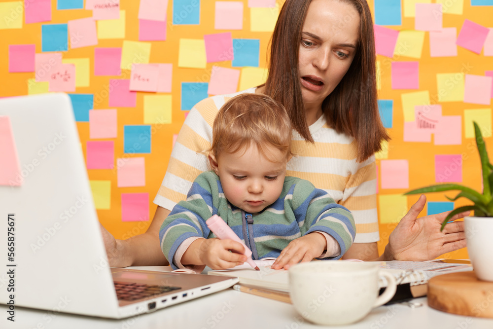 Portrait of shocked woman with brown hair wearing striped t-shirt sitting at table standing against yellow wall with colorful sticky notes, mother with kid in office, child messes up mommy's documents Portrait of shocked woman with brown hair wearing striped t-shirt sitting at table standing against yellow wall with colorful sticky notes, mother with kid in office, child messes up mommy's documents