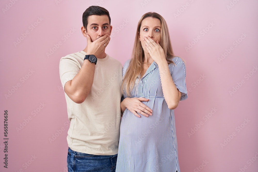 Young couple expecting a baby standing over pink background shocked covering mouth with hands for mistake. secret concept. Young couple expecting a baby standing over pink background shocked covering mouth with hands for mistake. secret concept.