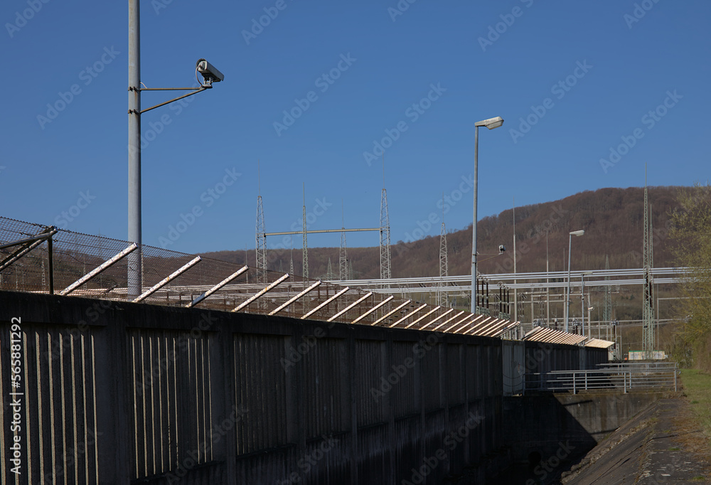 Würgassen, Germany - 03 23 2020: Surveillance camera on a pole behind a concrete and barbed wire fence. A substation is in the background. Würgassen, Germany - 03 23 2020: Surveillance camera on a pole behind a concrete and barbed wire fence. A substation is in the background.