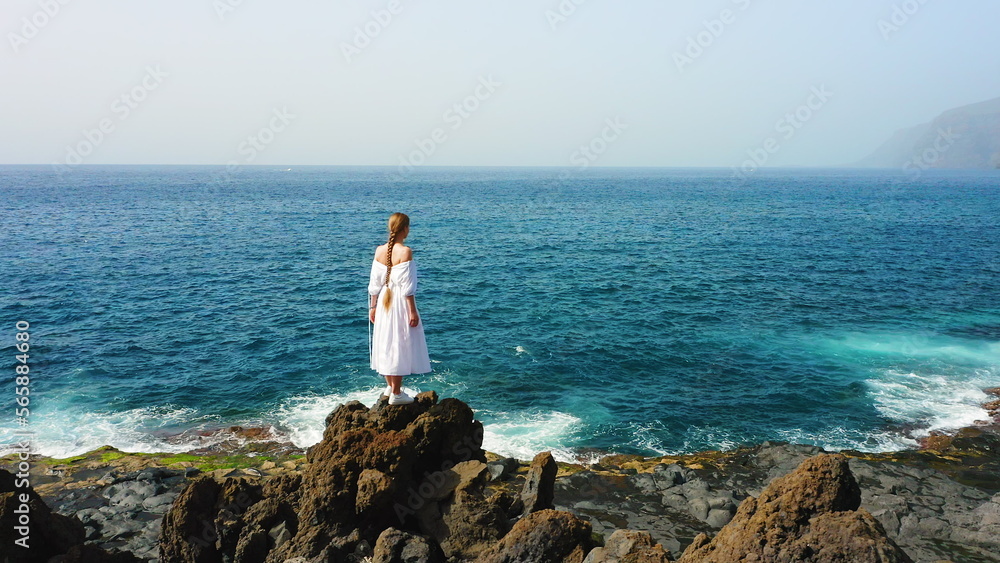 Female in white vintage dress on rocky volcanic beach.