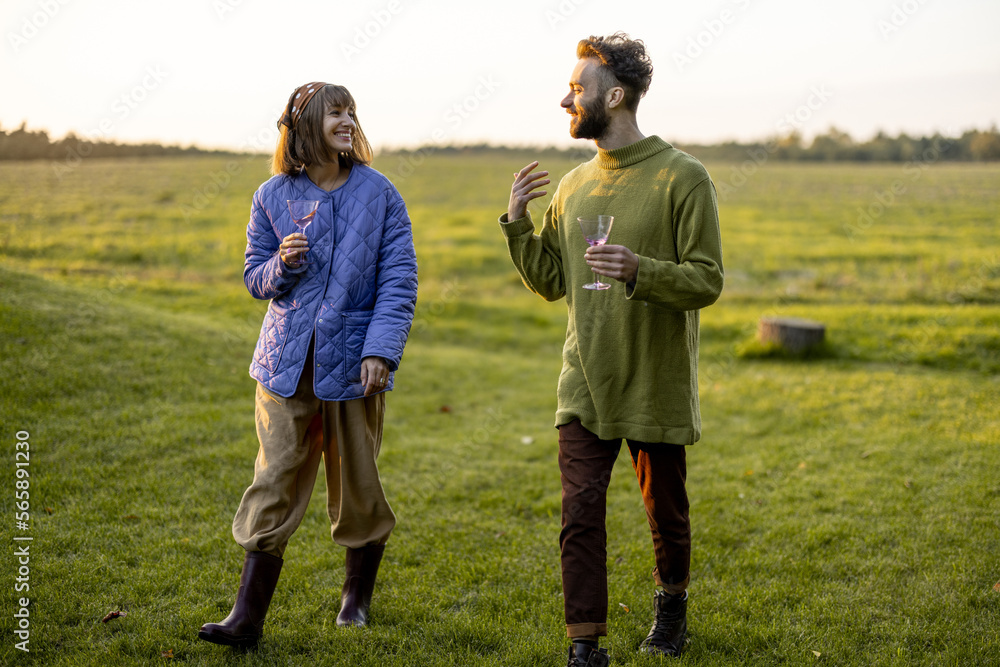 Stylish man and woman dressed warmly have conversation while walk together with wine on green lawn during a sunset. Couple spend leisure autumn time on nature