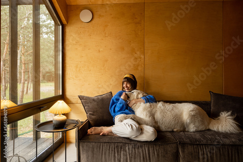 Woman cares her dog while sitting on a couch by the window, spending leisure time together in wooden house on nature