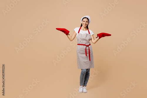 Full body smiling happy fun young housewife housekeeper chef baker latin woman wear apron toque hat spread hands in gloves potholders isolated on plain pastel light beige background ook food concept photo