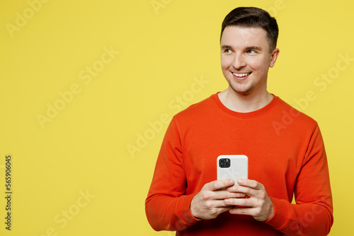 Young smiling cheerful happy man wear orange casual clothes hold in hand use mobile cell phone look aside on area isolated on plain yellow color background studio portrait. People lifestyle concept. © ViDi Studio