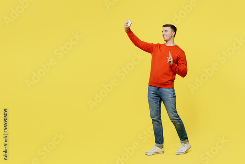 Full body young man wear orange casual clothes doing selfie shot on mobile cell phone post photo on social network isolated on plain yellow color background studio portrait. People lifestyle concept.