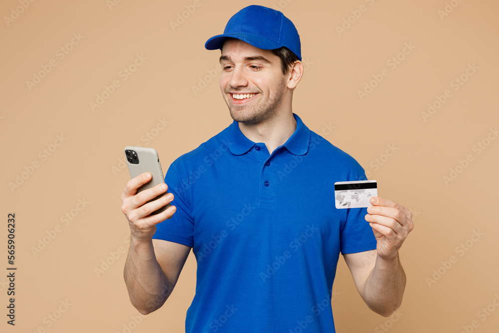Fun delivery guy employee man wear blue cap t-shirt uniform workwear work as dealer courier using mobile cell phone credit bank card isolated on plain light beige background. Online shopping concept. Fun delivery guy employee man wear blue cap t-shirt uniform workwear work as dealer courier using mobile cell phone credit bank card isolated on plain light beige background. Online shopping concept.