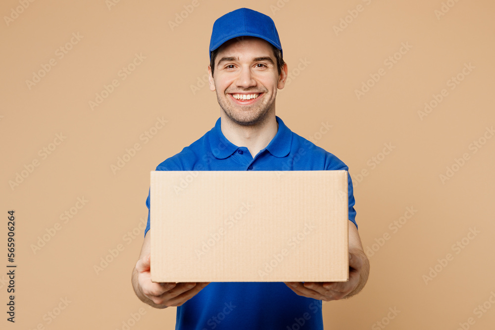 Professional smiling positive fun delivery guy employee man wear blue cap t-shirt uniform workwear work as dealer courier hold cardboard box isolated on plain light beige background. Service concept. Professional smiling positive fun delivery guy employee man wear blue cap t-shirt uniform workwear work as dealer courier hold cardboard box isolated on plain light beige background. Service concept.