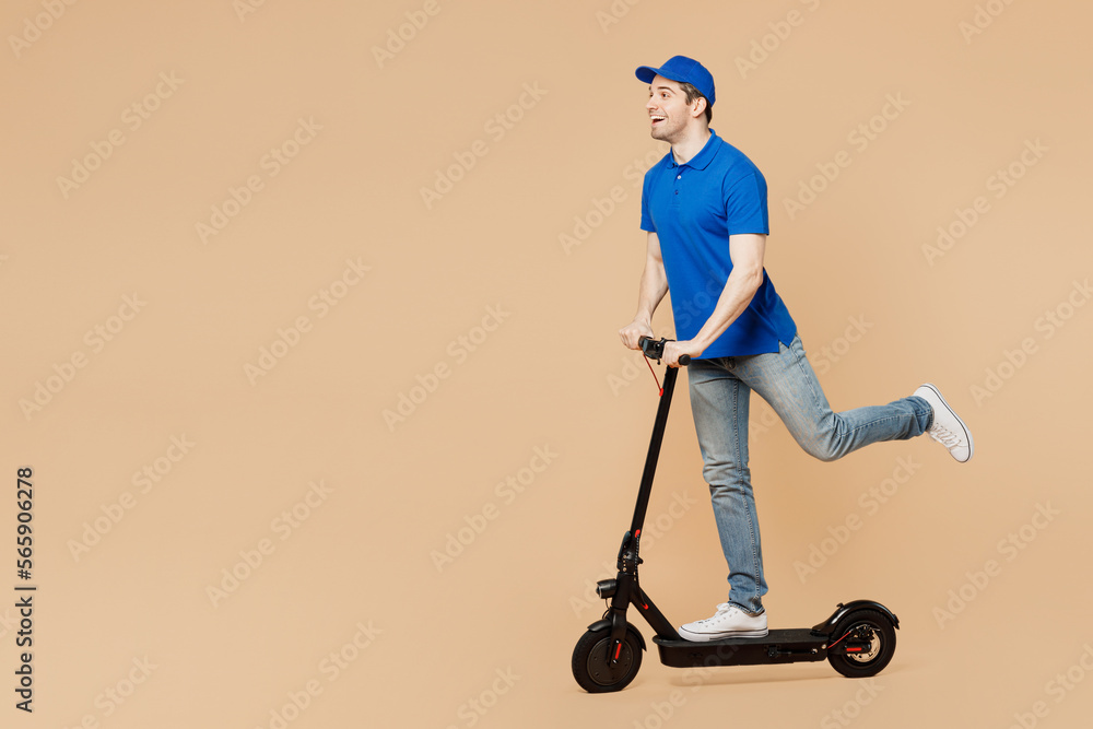 Full body side view happy delivery guy employee man wear blue cap t-shirt uniform workwear work as dealer courier riding e-scooter look aside isolated on plain light beige background. Service concept. Full body side view happy delivery guy employee man wear blue cap t-shirt uniform workwear work as dealer courier riding e-scooter look aside isolated on plain light beige background. Service concept.