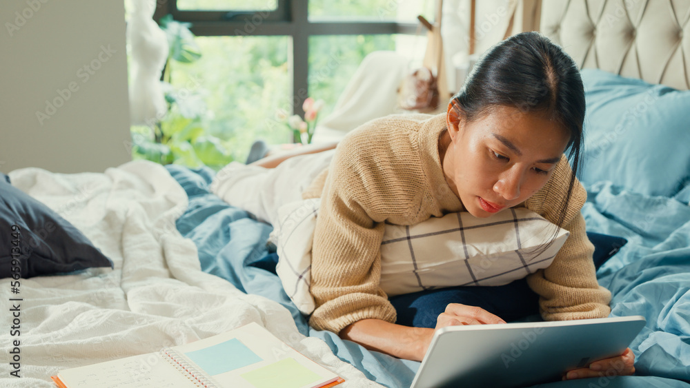 Happy Asian girl university with white cream pajamas lay down on fluffy comfort bed use digital tablet learning online in bedroom at home. Revisualizing flexible working, Work from home concept. Happy Asian girl university with white cream pajamas lay down on fluffy comfort bed use digital tablet learning online in bedroom at home. Revisualizing flexible working, Work from home concept.