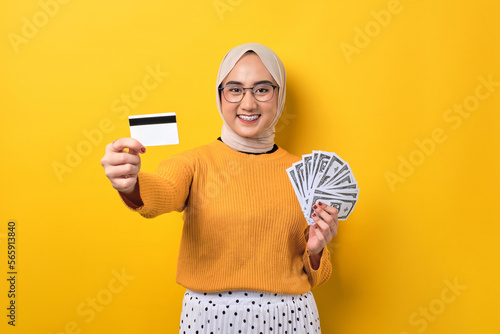 Beautiful cheerful Asian girl wearing hijab showing money banknotes and credit card isolated on yellow background