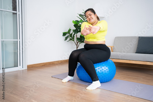 Portrait plus size woman doing exercise with fitness ball in home gym. Overweight woman sitting on a pilates ball and Stretching her muscles before exercising. Health care and weight loss concept.