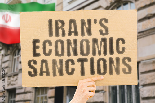 The phrase " Iran's Economic Sanctions " is on a banner in men's hands with blurred background. Manufacturing. Exportation. Import. Supply. Demand. Shortage. International. Diplomacy. Negotiation
