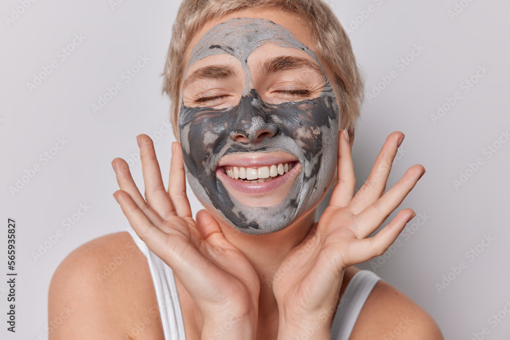 Close up shot of European woman smiles toothily applies nourishing clay mask undergoes beauty procedures wears t shirt stands bare shoulders isolated over grey background. Skin care treatment Close up shot of European woman smiles toothily applies nourishing clay mask undergoes beauty procedures wears t shirt stands bare shoulders isolated over grey background. Skin care treatment