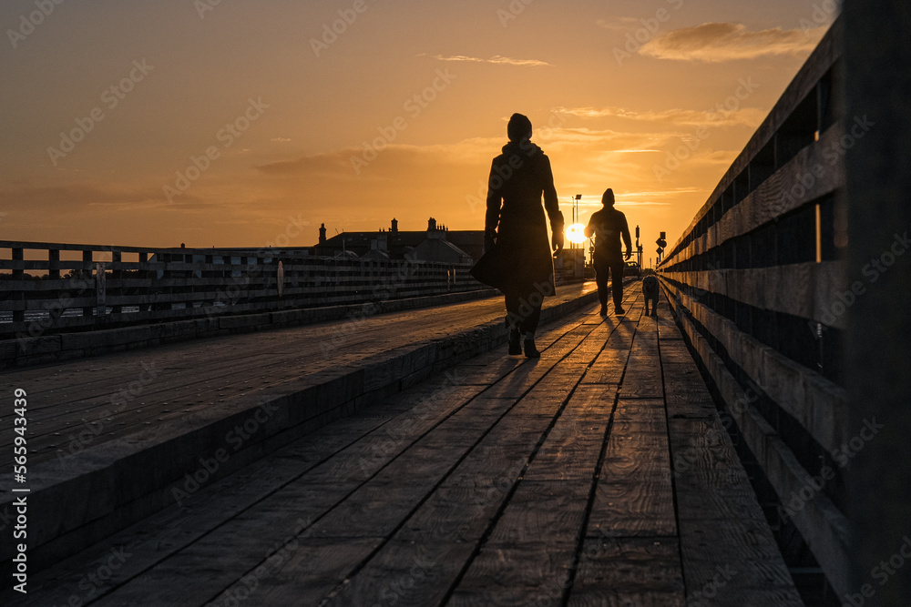 A woman, a men and a dog, walking on a wooden bridge during the sunrinse, with a clear sky
