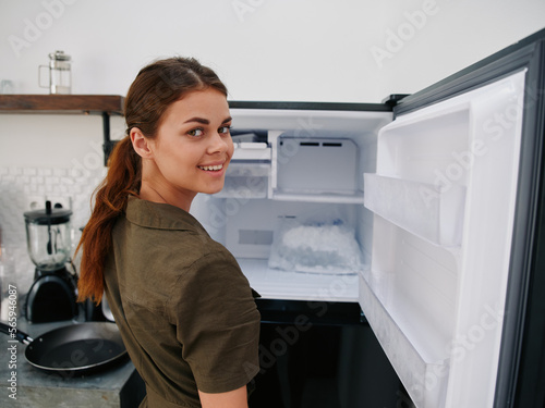Woman smiling with teeth looking into camera in kitchen at home opened freezer empty with ice inside, home refrigerator, defrosted, view from back, stylish interior.