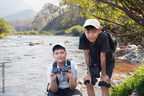 Young asian boy in plaid shirt and red cap holds binoculars in hands and watching the fish in the river during his vacation and his school project work in summer, soft focus, nature learning concept.