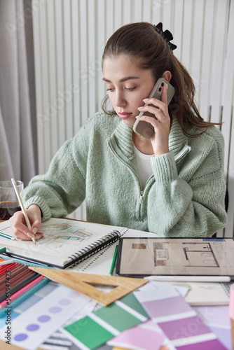 Creative femake designer works on apartment design draws sketches in spiral album surrounded by samples palette and stationery makes phone call to ask adive has serious concentrated expression photo