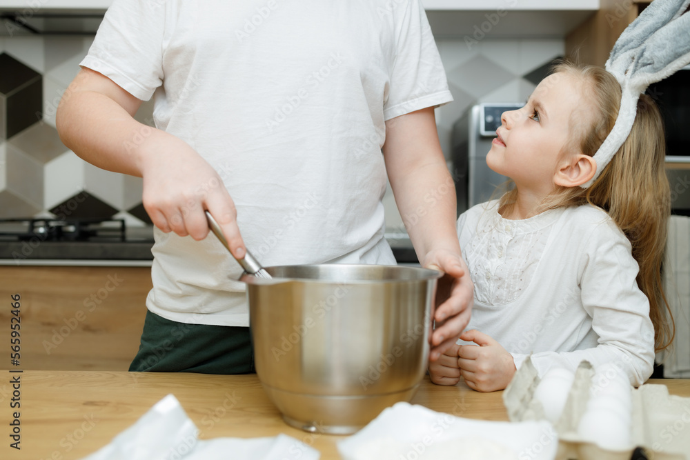 Siblings stand at kitchen table. Little girl in bunny ears looking at her big brother whisking dough in modern kitchen. Easter concept Siblings stand at kitchen table. Little girl in bunny ears looking at her big brother whisking dough in modern kitchen. Easter concept