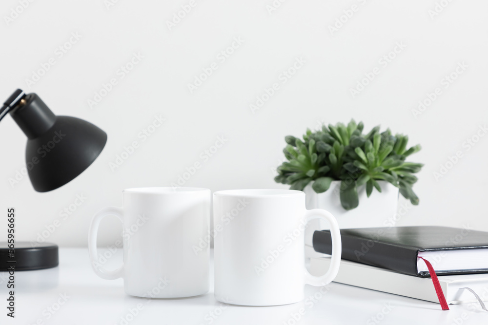 Two mugs mockup with notepad, accessories on white table and green plant in pot. Front view. Two mugs mockup with notepad, accessories on white table and green plant in pot. Front view.