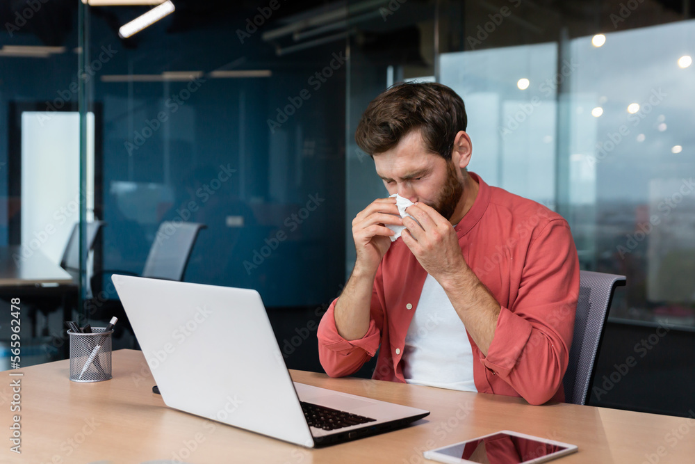 A young man is sick at work. sits at the desk in the office, wipes his nose with a napkin, has a runny nose, does not feel well. A young man is sick at work. sits at the desk in the office, wipes his nose with a napkin, has a runny nose, does not feel well.