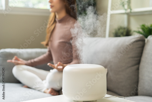 Air humidifier, calm blurred woman, girl sitting on couch lotus pose put hands practice meditation do yoga exercise at home. Aromatherapy steam scent from essential oil diffuser in living room at home photo