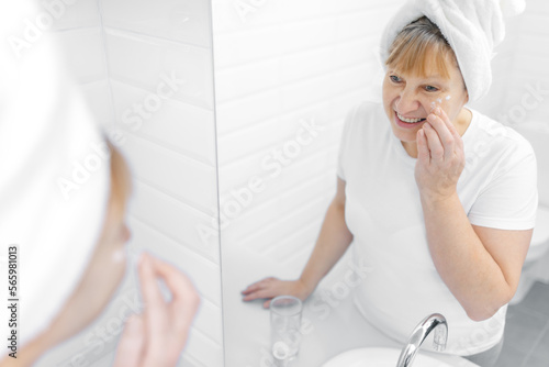 Gorgeous mid age older adult 50 years old blonde woman wears bathrobe in bathroom applying nourishing antiage face skin care cream treatment, looking at mirror doing daily morning beauty routine.