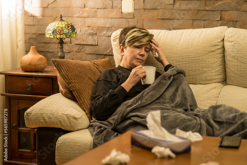 mujer mediana edad enferma con dolores  en el sofa de casa tapada con una manta, tomando medicina mirando el movil y el ordenador