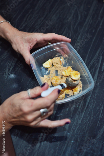 woman's hands eating sliced ​​banana with Peruvian maca oats in a clear pot with spoon on a dark marble table and table protector