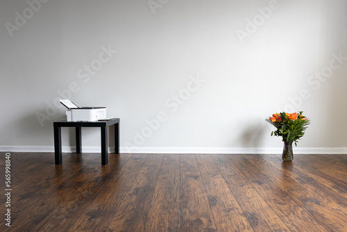 Fototapeta Naklejka Na Ścianę i Meble -  A room with nothing in it but a black table holding an inkjet printer and a vase filled with a roses floral arrangement on the dark laminated floor near the white wall's baseboard.