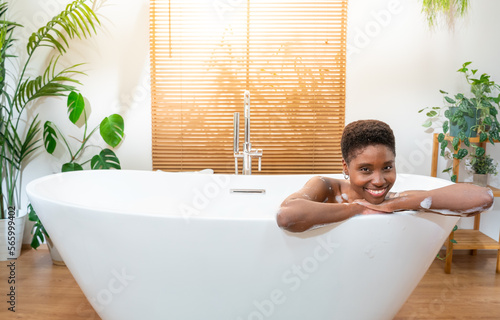 Luxury bath portrait of an attractive African woman relaxing in bathtub at cozy home bathroom looking at camera smiling happy. Real people wellness concept. High quality photo