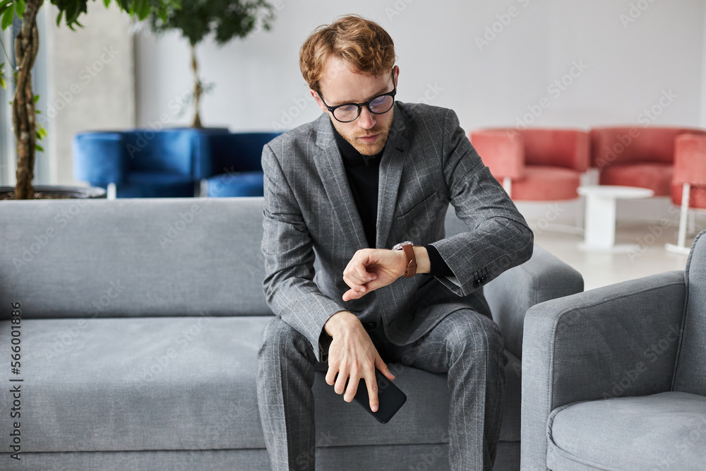 Male entrepreneur in suit, checking time on wrist watch, sitting in armchair in a recruitment agency Male entrepreneur in suit, checking time on wrist watch, sitting in armchair in a recruitment agency
