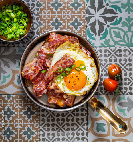 Fried eggs with bacon and green onions in a frying pan. Ethnic  tiles as background. Rustic breackfast. Top view.