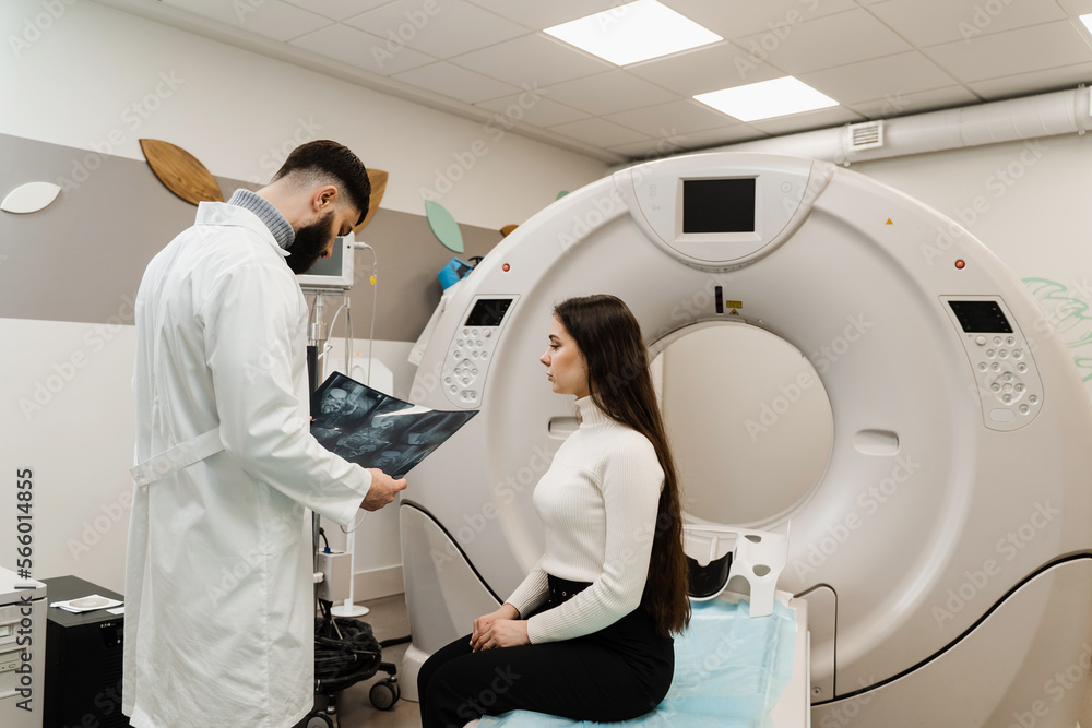 CT Doctor consulting patient and showing chest x-ray to patient in computed tomography room. CT scan radiologist showing x-ray of abdomen to girl patient in computed scanning room. CT Doctor consulting patient and showing chest x-ray to patient in computed tomography room. CT scan radiologist showing x-ray of abdomen to girl patient in computed scanning room.