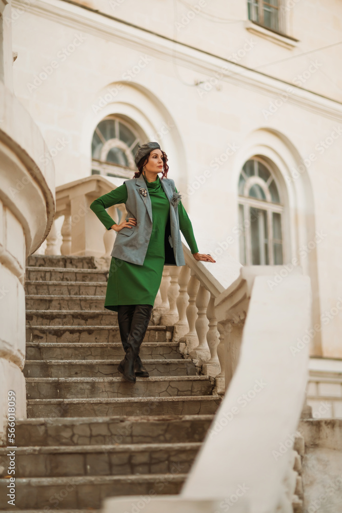 Outdoor fashion portrait of an elegant fashionable brunette model in a beret, green dress and a gray waistcoat posing at sunset in a european city in autumn. Outdoor fashion portrait of an elegant fashionable brunette model in a beret, green dress and a gray waistcoat posing at sunset in a european city in autumn.