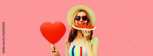 Portrait of happy young woman eating fresh watermelon with big red heart shaped balloon wearing summer straw hat, sunglasses on pink background © rohappy