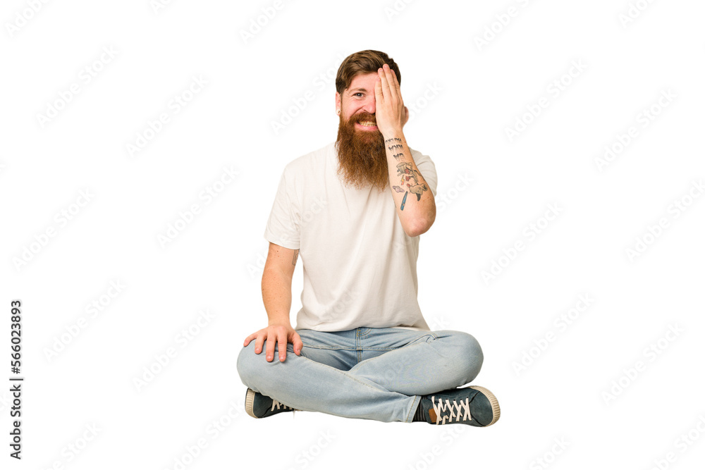 Young adult redhead with a long beard sitting on the floor isolated having fun covering half of face with palm.