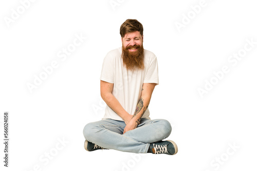 Young adult redhead with a long beard sitting on the floor isolated laughs and closes eyes, feels relaxed and happy.