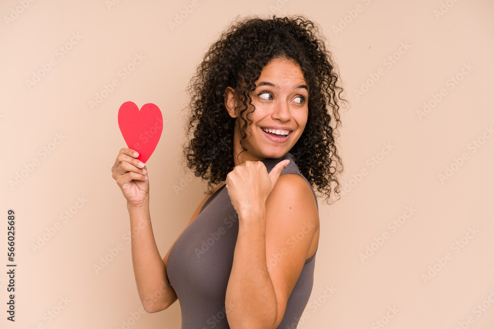Young african american woman holding a heart for valentines day isolated points with thumb finger away, laughing and carefree. Young african american woman holding a heart for valentines day isolated points with thumb finger away, laughing and carefree.