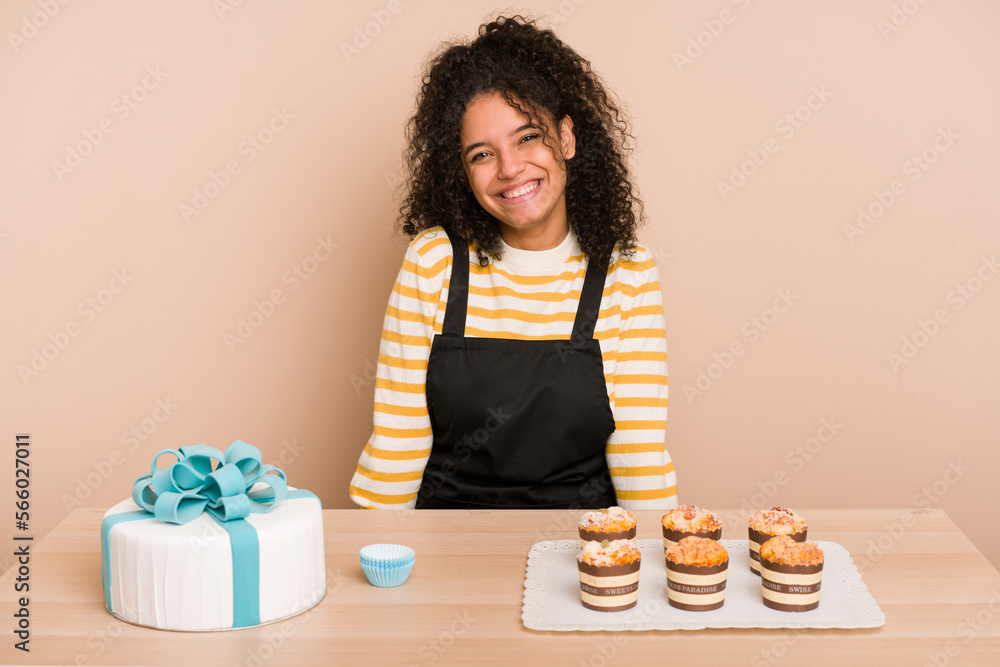 Young african american woman preparing a sweet cake and muffins on a table happy, smiling and cheerful. Young african american woman preparing a sweet cake and muffins on a table happy, smiling and cheerful.